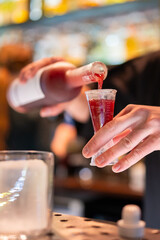 Bartender pouring red liquid from a bottle into a jigger at a bar. Close-up on hands and drink preparation with blurred lights in the background