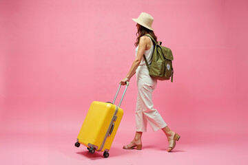 A full-length rear view of a female backpacker carrying a suitcase in motion against a vibrant, solid pink background.