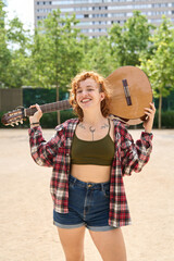 Portrait of smiling redhead young woman musician carrying acoustic guitar on shoulder in city park