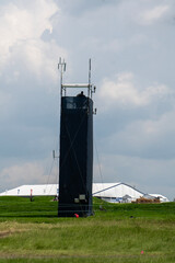  A tall, dark broadcast tower stands in a green field under a cloudy sky. A person is visible at...