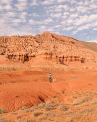 A woman stands with her hands raised in front of the beautiful red mountains of Bogutty her dog is next to her, they travel together through the unique nature of Kazakhstan