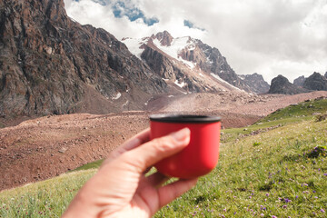 Female hand of tourist holding red thermos cup with hot drink against beautiful mountain landscape in summer