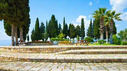 Public park with paved stone terraces, benches, picnic tables, and tall trees in Hierapolis near Pamukkale, Turkey. Sunny summer day with blue sky and scattered clouds in peaceful green surroundings.