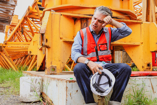Senior foreman overseeing large construction site sits down rest exhausted from hot weather thirsty dizzy headache fainting dehydrated wants drink water and suffers heatstroke due hot humid summer.