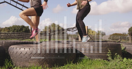 Hopping women in athletic wear over tractor tires on grassy field with wooden fence, monkey bars