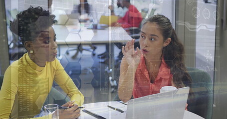 Women in yellow top and pink blouse collaborating at office table, with laptop and digital overlays