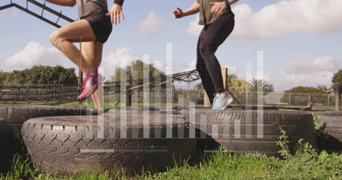 Hopping women in athletic wear over tractor tires on grassy field with wooden fence, monkey bars