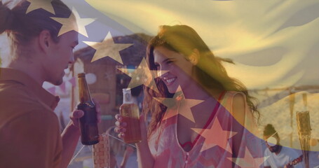 Toasting couple holding beer bottles clinking near beach cabana at sunset, with tiki torch