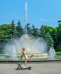 Happy young woman having fun with an electric scooter in a city park by a fountain on a sunny summer day.Urban mobility and summer lifestyle.Eco-friendly transport concept