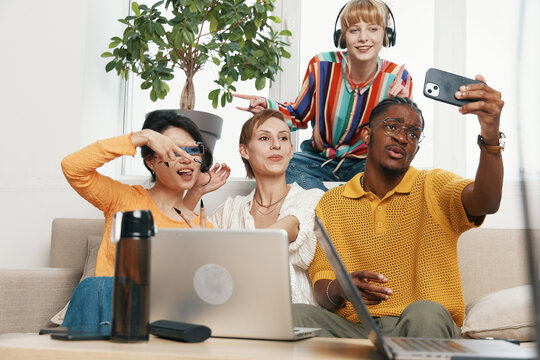 Young adult coworkers and friends socializing while working together in a modern workplace, capturing a fun moment with diverse Gen Z colleagues using laptops and smartphones.