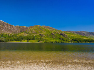 Clear lake with a wide gravel beach and green mountains under a bright summer sky (Buttermere, Lake District, England, United Kingdom)