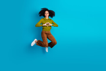 Joyful young woman jumping with heart hand gesture against a vibrant blue background in fashionable...