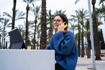 Young woman attending online classes or smart working using digital tablet and smartphone outdoors in a park with palm trees