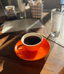 Coffee cup filled with dark brew sits on an orange saucer, surrounded by glassware and a menu, illuminated by warm sunlight creating a cozy atmosphere