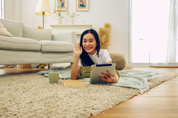 Beautiful asian woman living free life waving hello lyingcarpet playing tablet with both legs...
