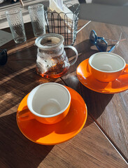 Two empty orange coffee cups on saucers sit on a wooden table beside a glass teapot filled with tea, creating a cozy cafe atmosphere with sunlight streaming in