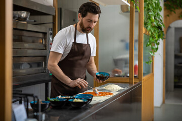 Male cooker working in the pizzeria kitchen and putting topping to the pizza