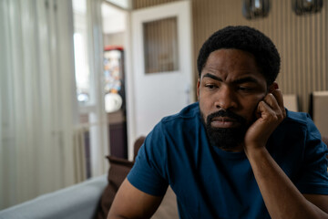 Depressed young black man sitting indoors looking down, feeling sad and stressed, emotional...