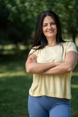 Smiling beautiful woman standing with crossed arms in park wearing sportswear, enjoying fresh...