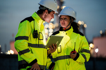 Engineer worker watching work monitor on digital tablet and checking for quality control at construction site of petroleum chemical industry or refinery oil plant factory overtime at night
