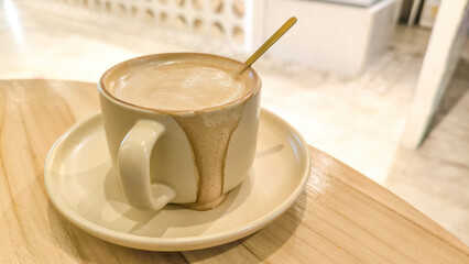 A ceramic cup filled with creamy coffee on a saucer with a golden spoon, placed on a wooden table in a bright, modern cafe setting.