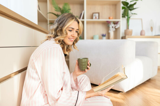 A relaxed woman reads on the floor with a cup of tea, surrounded by tranquility and the comforts of her inviting home.