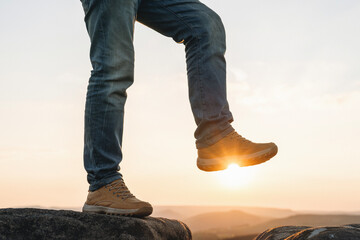 Person hiking on rocky terrain during sunset with sun rays peeking through footwear, showcasing...