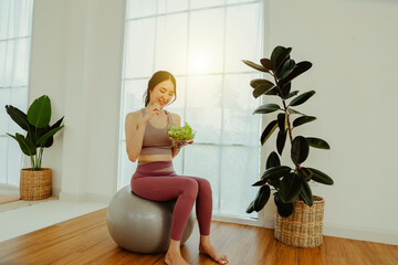 Happy Asian woman in yoga exercise clothes sitting on yoga ball smiling holding salad bowl and eating fresh salad. Fat free food concept. Healthy diet food and eating healthy.