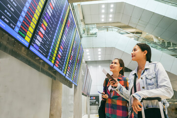 Two female asian passengers who were friends holding tickets for an international trip stood in the departure terminal and looked at the departure board with interest to check their own flights.