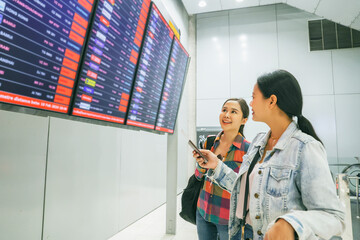 Two female asian passengers who were friends holding tickets for an international trip stood in the departure terminal and looked at the departure board with interest to check their own flights.