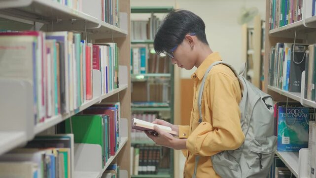 Waist up medium shot of male student walking in college library and taking textbook from shelf. Male Asian student browsing through book in background.