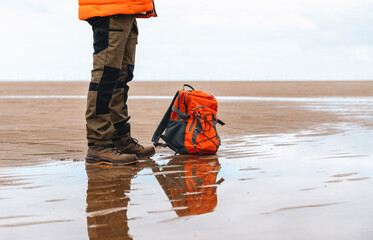 Person standing on wet sand at the beach with an orange backpack during overcast weather in late...
