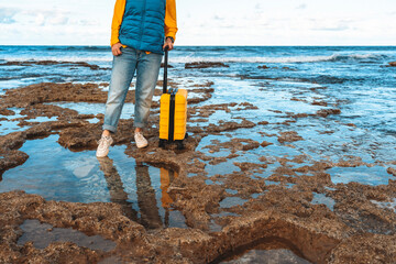 Traveler explores rocky beach at low tide while pulling yellow suitcase under bright sky and calm...
