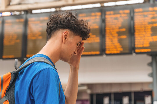 Young traveler checking train schedules at busy transit station during the day while looking concerned about delays and connections