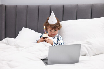 Birthday celebration. Little boy with cupcake having video call via laptop in bed at home