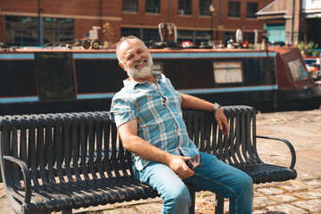 Senior man relaxing on park bench near canal boat in urban setting during sunny daytime; enjoying...