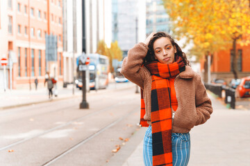 Young woman in cozy autumn attire poses on city street with orange and yellow leaves in the...