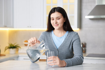 Woman pouring water into glass at white marble table in kitchen