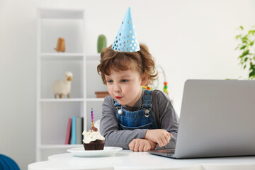 Birthday celebration. Little boy blowing candle on cupcake while having video call via laptop at...