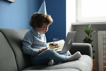 Birthday celebration. Little boy blowing candle on piece of cake while having video call via laptop...