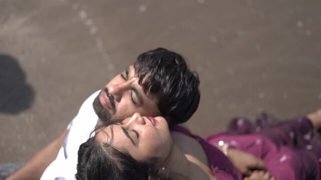 A cheerful Indian couple enjoys a sunny day at the beach, holding hands while walking along the shoreline. The woman is dressed in a casual maroon dress, and the man wears a white t-shirt with grey sh