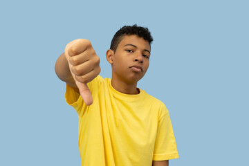 Dark skinned Latino teenager wearing a yellow shirt with a disappointed face making a thumbs-down...