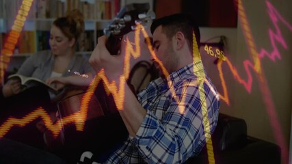 Man guitarist reacting to financial graph fluctuations by strumming chord progressions on couch