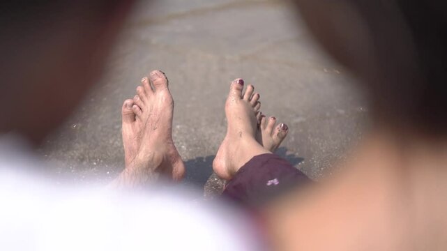 A cheerful Indian couple enjoys a sunny day at the beach, holding hands while walking along the shoreline. The woman is dressed in a casual maroon dress, and the man wears a white t-shirt with grey sh