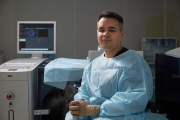 Portrait of man sitting in operating room wearing surgical gown and gloves, looking at camera with...