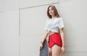 Stylish young woman holding a longboard, leaning against a textured wall. Modern skate culture and fashion.