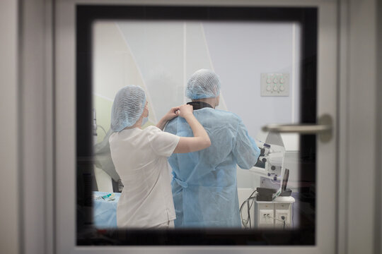 woman assisting man by fastening surgical gown in operating room, both wearing disposable caps and standing near medical equipment