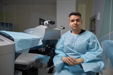 Portrait of man sitting in operating room wearing surgical gown and gloves, looking into camera...