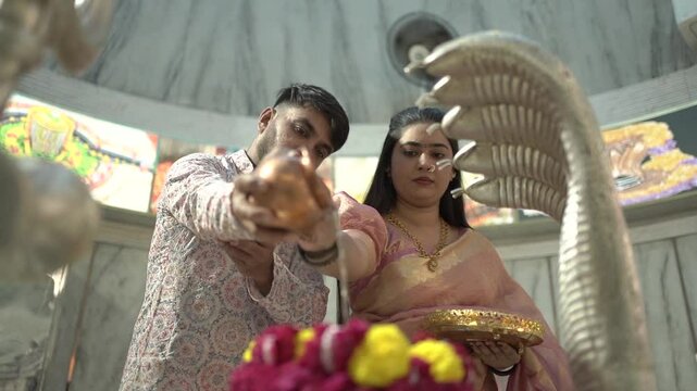 An Indian couple performs the sacred Abhishekam ritual to a Shiva Lingam in a Hindu temple. The woman is dressed in an elegant silk saree with jasmine flowers in her hair, and the man wears traditiona