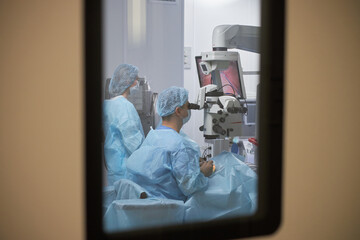 Two women in surgical gowns performing operation using advanced medical microscope in modern...